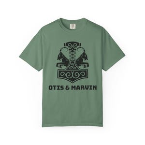 Puede incluir: Camiseta verde salvia con un diseño negro de un martillo con dos carneros y el texto "OTIS & MARVIN". La camiseta tiene cuello redondo y mangas cortas. El diseño está centrado.