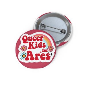 May include: Two round, red and white pinback buttons. One button features the text "Queer Kids for Ares" in red with a rainbow and flower graphic. The other button is red and white striped.