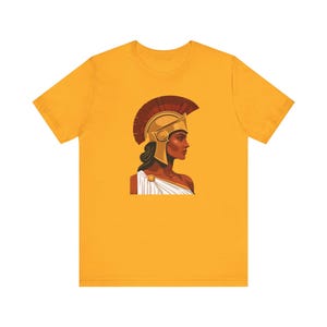 Puede incluir: Camiseta de color amarillo mostaza con el perfil de una mujer con un casco dorado y una cresta roja. La mujer lleva una toga blanca. El gráfico es una ilustración estilizada.