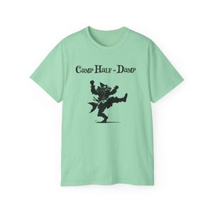 May include: A light green t-shirt featuring the text "Camp Half-Damp" above a black illustration of a dancing creature. The creature is wearing a hat and a sash. The t-shirt has short sleeves and a crew neck.