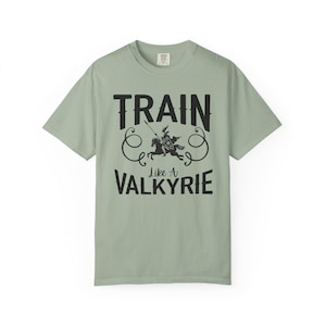 May include: Sage green t-shirt with the text "TRAIN Like A VALKYRIE" in black. The design includes a graphic of a warrior on horseback with a spear. The shirt is a short-sleeved crew neck.