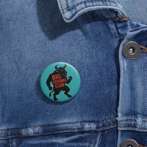 Pode incluir: Um botão redondo, turquesa com o texto "BIG FEELINGS CLUB" em preto e vermelho. O botão apresenta uma silhueta preta de um touro. O botão está preso a uma jaqueta jeans azul.