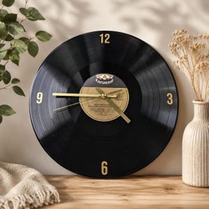 Op de afbeelding: Een ronde wandklok gemaakt van een zwarte vinylplaat met goudkleurige cijfers op de posities 12, 3, 6 en 9 uur. Het centrale label toont het Karussell-logo en tekst, voorzien van dunne goudkleurige wijzers voor de uren, minuten en seconden.