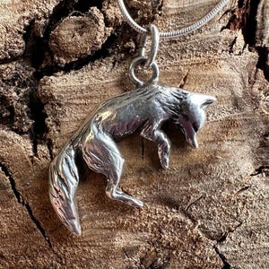 May include: A silver pendant necklace featuring a detailed fox charm. The fox is depicted in a downward pose, with its tail curved. The pendant hangs from a delicate silver chain. The background is a textured, brown wooden surface.