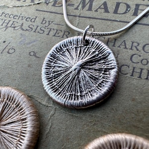 Peut inclure: Un collier avec un pendentif en argent de forme circulaire qui ressemble à un bouton tissé. Le pendentif est sur une chaîne en argent et repose sur un morceau de papier avec du texte.