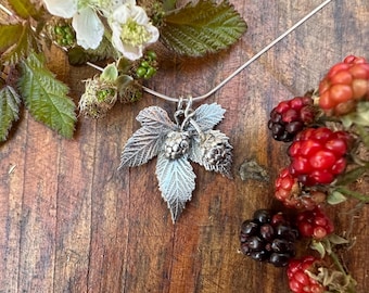 May include: A silver pendant featuring a detailed blackberry leaf and two small blackberry fruits, suspended from a thin silver chain. The metal surface shows intricate textures and veins, capturing the natural form of the plant in metallic detail.