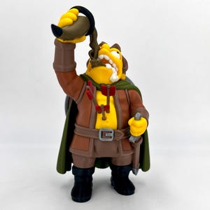 May include: A yellow-skinned character figurine wearing a brown tunic, green cape, and black boots. The figure holds a dark drinking horn overhead, with liquid pouring into its mouth. Two red-fletched arrows are embedded in the chest, and a silver-colored dagger hangs from a belt at the waist.