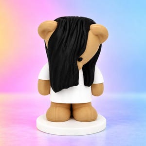 Puede incluir: Una figura de oso color canela sobre una base circular blanca. El oso tiene el cabello negro, largo y liso, cubriendo su rostro y viste una camiseta blanca lisa de manga corta. La figura tiene un acabado suave y mate.
