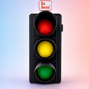 Puede incluir: Un semáforo negro con tres luces circulares en rojo, amarillo y verde. Un pequeño letrero rectangular se sitúa sobre el semáforo con un borde rojo y el texto Go Away! impreso en letras rojas sobre un fondo de color blanco.