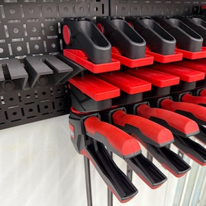 May include: A wall-mounted storage system for holding multiple black and red clamp-style tools. The tools are arranged in rows, with the handles facing outward.
