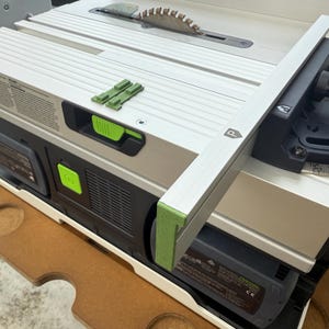 May include: A close-up of a table saw with a green and silver fence attached. The fence has a level and a green rubber strip along the edge. The saw has a black and green power switch.