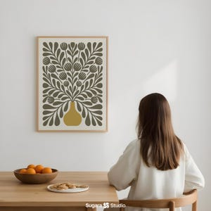 May include: A framed art print featuring a floral design in olive green and mustard yellow. The artwork depicts a vase filled with stylized flowers and foliage. The print is displayed on a white wall.