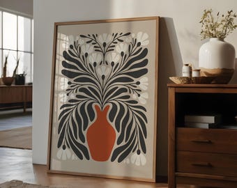Op de afbeelding: Een ingelijste kunstdruk met een gestileerd bloemmotief. Het kunstwerk toont een terracotta vaas gevuld met zwart en wit blad tegen een neutrale achtergrond. De print zit in een houten lijst, wat een vleugje warmte aan het decor toevoegt.