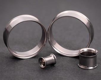 May include: Four silver metal ear gauges of different sizes. The gauges are circular and have a smooth, polished finish.