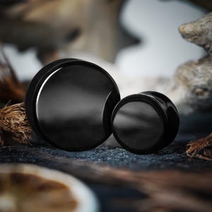 Puede incluir: Dos tapones para los oídos negros de diferentes tamaños. Los tapones tienen un acabado liso y brillante. El tapón más grande tiene aproximadamente 2,5 cm de diámetro, y el tapón más pequeño tiene aproximadamente 1,3 cm de diámetro. Están colocados sobre un fondo oscuro y texturizado con elementos naturales.