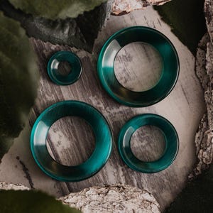 Puede incluir: Cuatro expansores circulares para orejas de color turquesa de varios tamaños están dispuestos sobre una superficie gris texturizada. Los expansores tienen un acabado brillante y un aspecto veteado. El más pequeño es un disco pequeño, los otros son anillos más grandes.