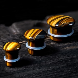 May include: Three tiger's eye stone plugs in different sizes. The plugs feature a rounded top with brown and gold bands. Each plug has a clear, rubber O-ring. They are displayed on a dark wooden surface.