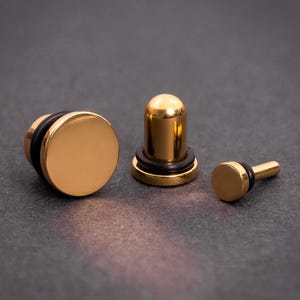 Könnte beinhalten: Drei goldfarbene Ohrstecker, einer mit runder Fläche, einer mit zylindrischer Fläche und einer mit kleiner runder Fläche und einem Stift.