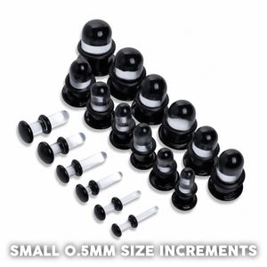 Puede incluir: Un surtido de tapones para los oídos en blanco y negro y medidores de oídos transparentes. Los tapones para los oídos tienen forma redondeada con un disco plano en cada extremo. Los medidores son transparentes con discos negros. El texto "SMALL 0.5MM SIZE INCREMENTS" está en la parte inferior.