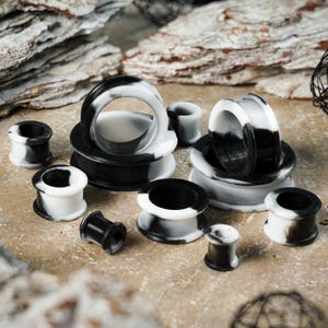 May include: A selection of black and white marbled ear gauges in different sizes. The gauges are made from a smooth material and have a circular design. Some are stacked, while others are displayed individually. The background has a textured, neutral surface.