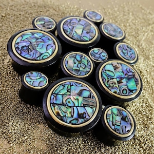 Puede incluir: Un conjunto de tapones para los oídos negros con incrustaciones iridiscentes de concha de abulón. Cada tapón tiene un borde dorado. Los tapones, de varios tamaños, están dispuestos sobre una superficie dorada brillante. La concha de abulón muestra tonos azules, verdes y morados.