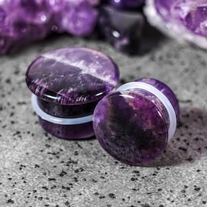Puede incluir: Un par de dilatadores de oreja de piedra amatista con un respaldo blanco. Los dilatadores son redondos y tienen un patrón de color morado y blanco.
