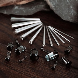 May include: An assortment of body jewelry, including silver-colored tapers and black and clear plugs, arranged on a dark wood surface. The tapers are cylindrical and pointed, while the plugs have a rounded, flared design.