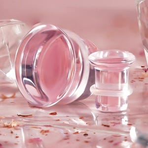 May include: Clear, pink-tinted glass ear gauges. One gauge is lying on its side, revealing its pink interior. Another gauge stands upright, showcasing its cylindrical design. The background is a soft pink, with scattered floral accents.