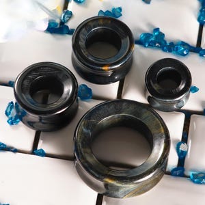 May include: Four dark blue and gold stone ear gauges of varying sizes are displayed on a white tiled surface. The gauges have a smooth, polished finish and a circular design. Blue decorative crystals surround the gauges.
