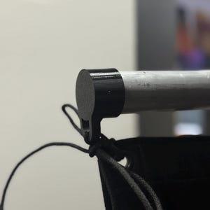 May include: A black plastic end cap attached to a silver metal rod. The cap features a small downward-facing hook with a thin black string tied to it. The string connects to a black fabric panel hanging vertically below the rod.