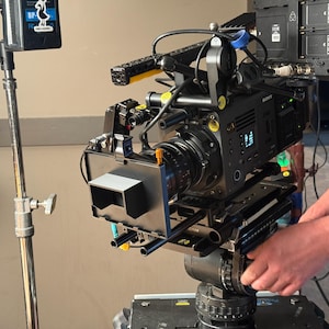 Może przedstawiać: Profesjonalna czarna kamera wideo zamontowana na statywie. Kamera posiada różne akcesoria, w tym matte box i monitor. Kamera jest regulowana przez osobę. Tło to neutralna ściana.