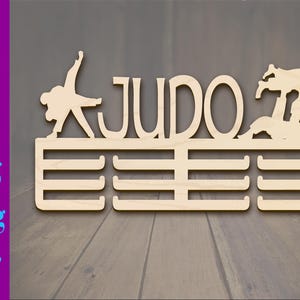 Puede incluir: Expositor de medallas de madera con la palabra "JUDO" en letras grandes y dos siluetas de atletas de judo. El expositor tiene cinco ranuras para medallas.