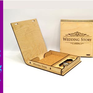 Puede incluir: Una caja de madera con una tapa con bisagras y un hueco para un objeto pequeño. La caja está decorada con las palabras "Wedding Story" y un diseño decorativo. La caja está abierta y la tapa está levantada.