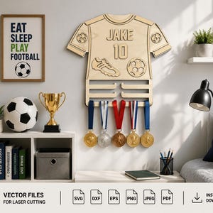 May include: A light-colored wooden wall-mounted medal hanger shaped like a soccer jersey, featuring the name JAKE and the number 10 engraved on the front. The design includes a soccer ball and cleat graphic, with five horizontal bars for hanging medals.