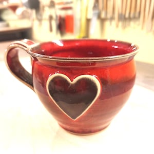 Könnte beinhalten: Handgefertigte Keramik-Tasse in tiefroter Farbe mit herzförmigem Ausschnitt. Die Tasse hat einen braunen Henkel und Rand. Der Herzausschnitt ist mit dunklerem Braun gefüllt.