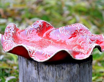 Puede incluir: Un plato de cerámica rojo con un diseño floral blanco. El plato tiene un borde ondulado y está sentado sobre un tocón de árbol.