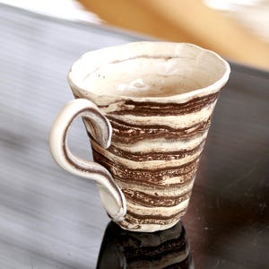 May include: A white ceramic mug with brown stripes. The mug has a unique, curved handle.