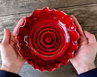 Puede incluir: Un cuenco de cerámica rojo vibrante con un borde ondulado y un ribete dorado. El interior del cuenco presenta un diseño en espiral en tonos de rojo, negro y marrón. El cuenco se sostiene con dos manos, mostrando su forma y color.