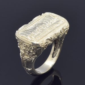 Può includere: Anello color argento con una superficie rettangolare con un design a rilievo dettagliato. L'anello presenta dettagli ornati, simili a fiori, sui lati e una croce sulla fascia. L'anello è probabilmente in argento sterling.