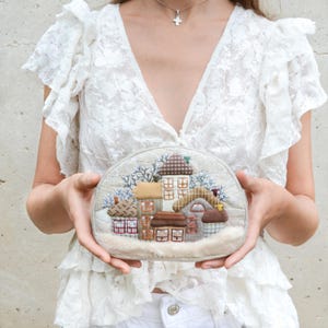 May include: A beige, half-moon shaped clutch featuring a detailed, fabric-appliqued village scene. The scene includes several small houses with brown, tan, and green roofs, surrounded by blue and white accents, and a faux fur trim.