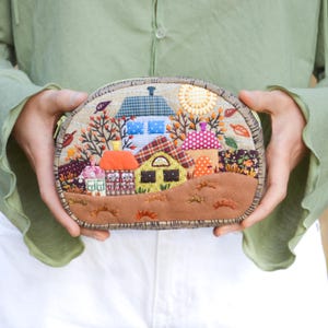 Op de afbeelding: Een ronde stoffen etui met een grillige dorpsscène. Het ontwerp toont kleurrijke huizen, bomen en een zon, allemaal gestikt op een beige achtergrond. De etui heeft een bruine suède-achtige basis en een ritssluiting.