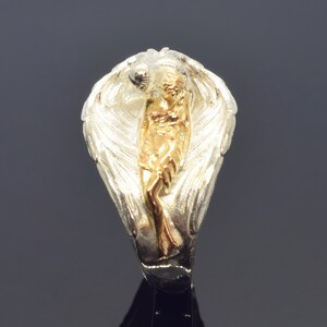 A silver ring with a gold-colored figure of a woman on it, set against a dark background.