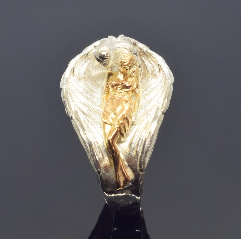 A silver ring with a gold-colored figure of a woman on it, set against a dark background.