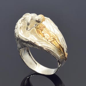 A silver ring with a gold-colored female figure on it, set against a dark background.