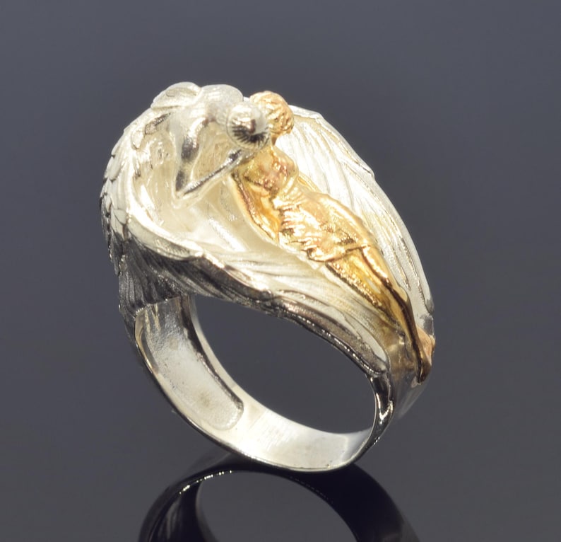 A silver ring with a gold-colored female figure on it, set against a dark background.
