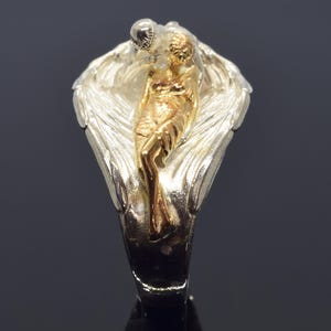 A close-up image of a ring featuring a gold-toned figure of a woman with a winged design.
