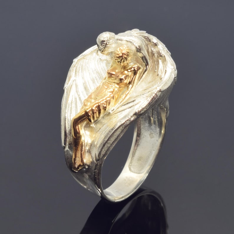 A silver ring with a gold-colored figure of a person sitting on a wing, which appears to be a decorative piece of jewelry.