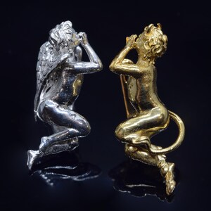 Könnte beinhalten: Zwei kniende figürliche Broschen. Eine ist silberfarben mit Flügeln, die andere goldfarben mit einem Schwanz. Beide Figuren knien, die Hände zum Gesicht erhoben. Die Broschen sind vor einem dunklen Hintergrund platziert.