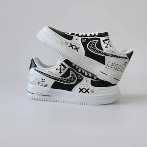 May include: A pair of black and white sneakers featuring a graphic design with the word LEGEND, various symbols, and cross patterns. The shoes have white laces, a white midsole with AIR text, and a black and white checkered swoosh logo on the side.