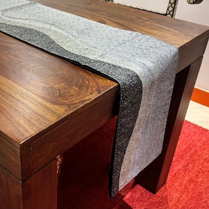 Può includere: Un runner da tavolo decorativo con un design strutturato a strati nei toni del grigio e del nero. Il runner è drappeggiato su un tavolo di legno scuro, aggiungendo un tocco di eleganza alla zona pranzo. Le dimensioni non sono visibili.